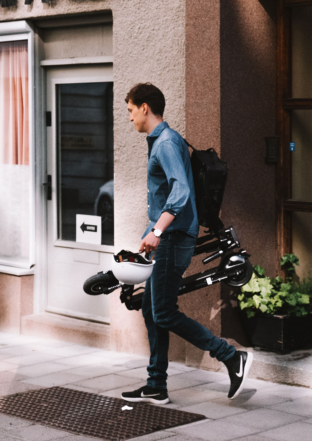 Man walks out the door holding a folded elecetric scooter and a white helmet