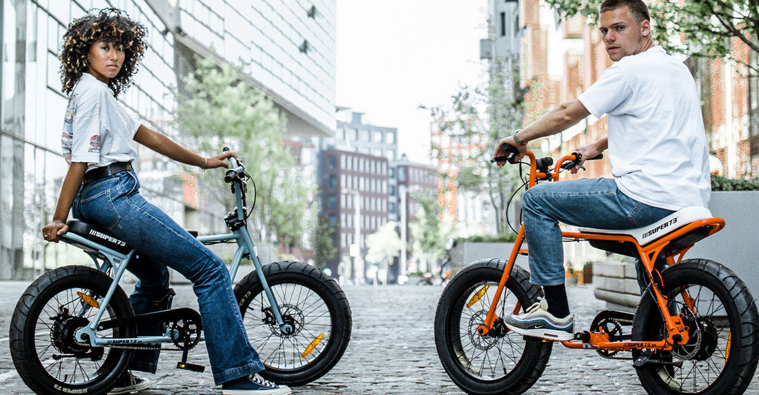 A boy and a girl sitting on two electric bikes looking in to the camera in front of big buildings