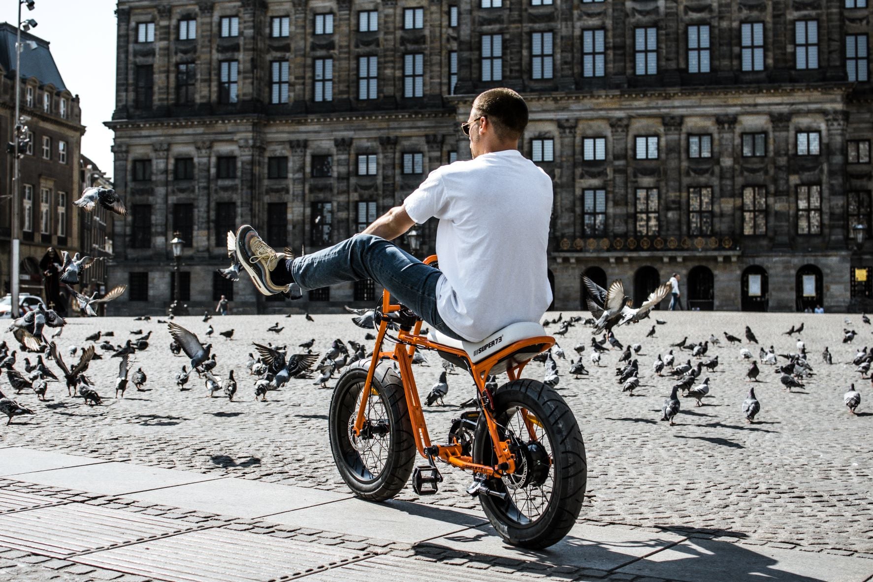 Man riding an super73 electric bike on the square with his feet in the air and a lot of birds flying around him