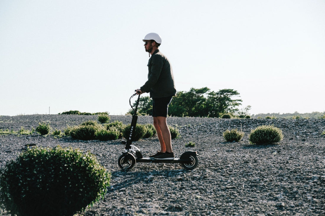 man rides an electric scooter on a rocky beach wearing a white helmet